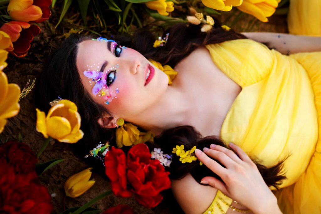 A woman in a bright yellow dress lies on the ground among red and yellow tulips, with flowers in her hair and decorative floral makeup on her face, creating a dreamy, fairytale-like scene.