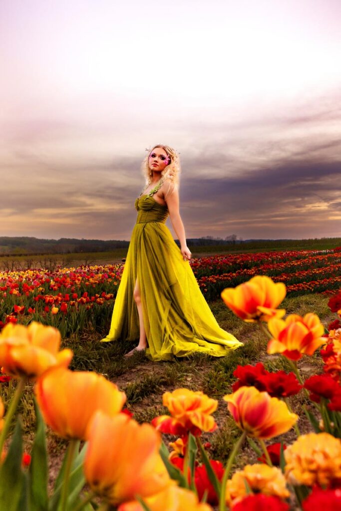 A woman in a long, flowing green dress sits on the edge of a white bathtub placed in a field of colorful tulips at sunset. She has wavy blonde hair, floral accessories, and looks off into the distance.