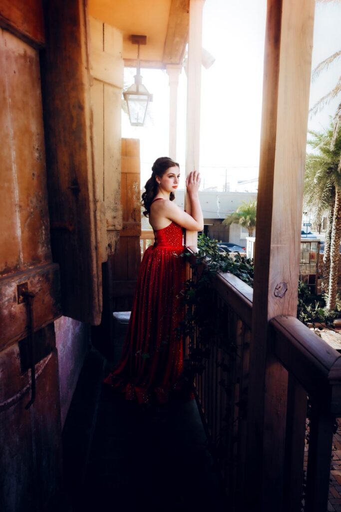 A woman in a red, strapless gown stands on a wooden balcony, gazing outward. Sunlight filters through, illuminating her and the tropical plants below.