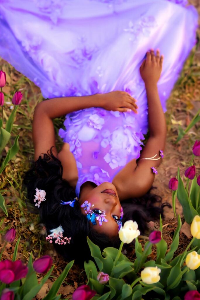 A woman with long dark hair lies amid flowers, wearing a purple dress with 3D floral designs. Her makeup features vivid purple and blue butterflies and petals on her face, creating a whimsical, enchanting look.