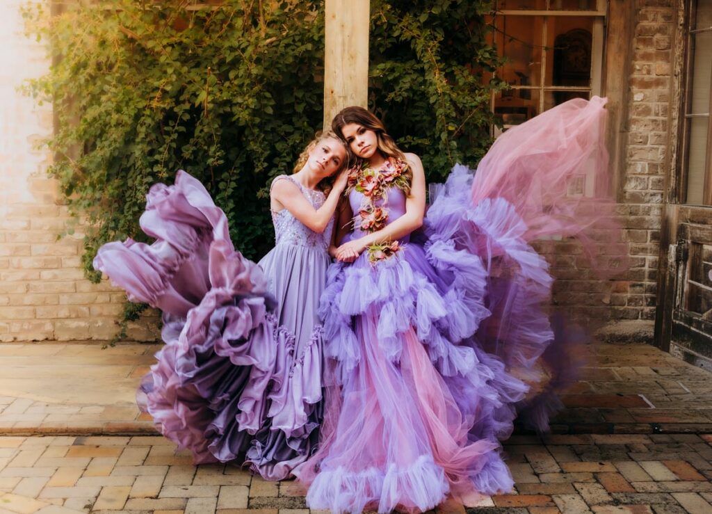 Two women in elaborate, ruffled lavender and purple gowns pose closely together outdoors, against a backdrop of ivy-covered brick walls and rustic windows. The dresses flow dramatically around them.