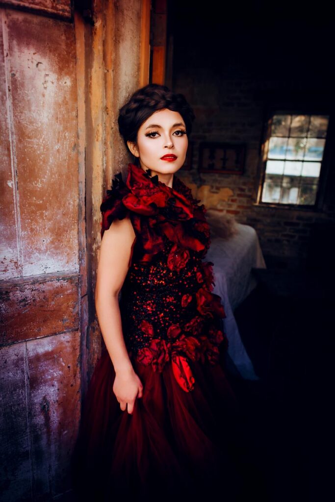 A woman in a dramatic, dark red gown with floral details stands against a rustic wooden door in a dimly lit room, gazing confidently at the camera. The background includes a brick wall and a partially visible window.