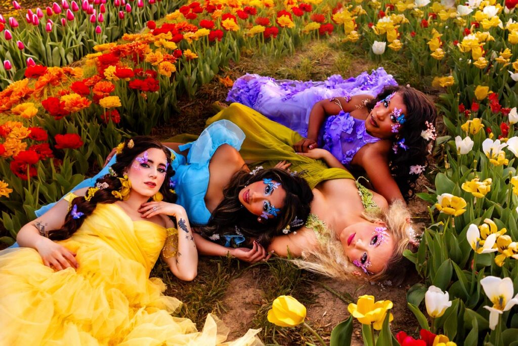 Four women in colorful, flowing dresses lie on the ground among vibrant red, yellow, and white tulips. Their faces are adorned with glitter and floral decorations, creating an ethereal, fairy-tale atmosphere.