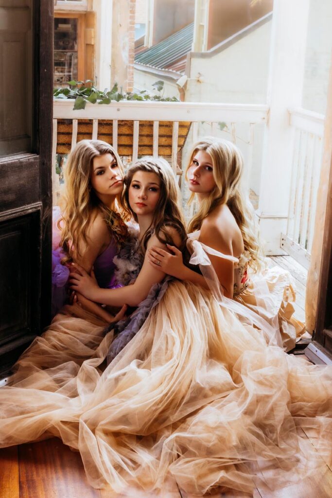 Three young women with long, wavy hair sit closely together on a wooden floor, wearing flowing, pastel-colored gowns. Soft light streams through a nearby window, creating a dreamy, ethereal atmosphere.