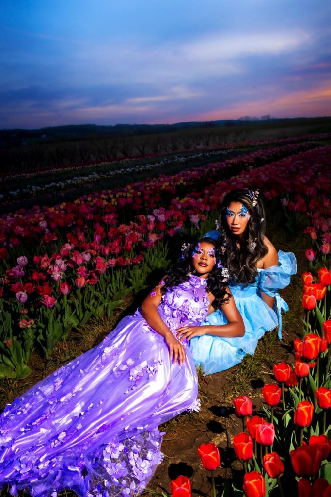 Two women in bright gowns, one in purple and one in blue, pose among vibrant red and pink tulips at sunset. They wear floral accessories and dramatic makeup, creating a dreamy, enchanted atmosphere in the flower field.