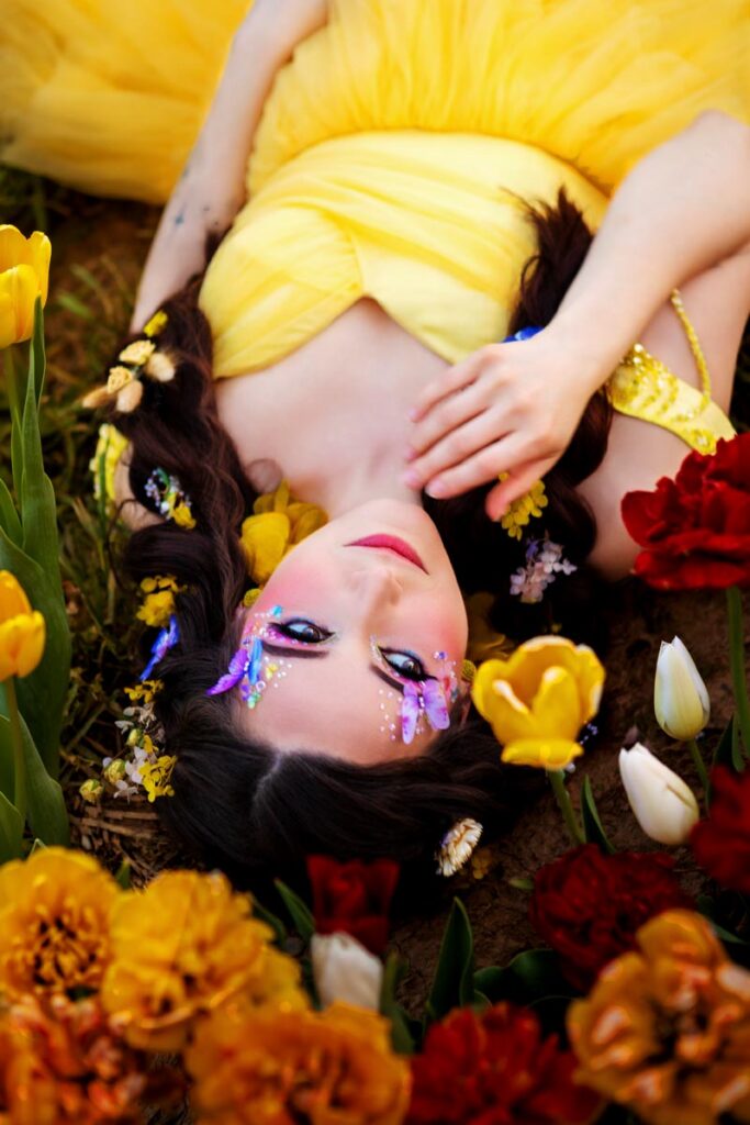 A woman in a bright yellow dress lies on the ground among red and yellow tulips, with flowers in her hair and decorative floral makeup on her face, creating a dreamy, fairytale-like scene.