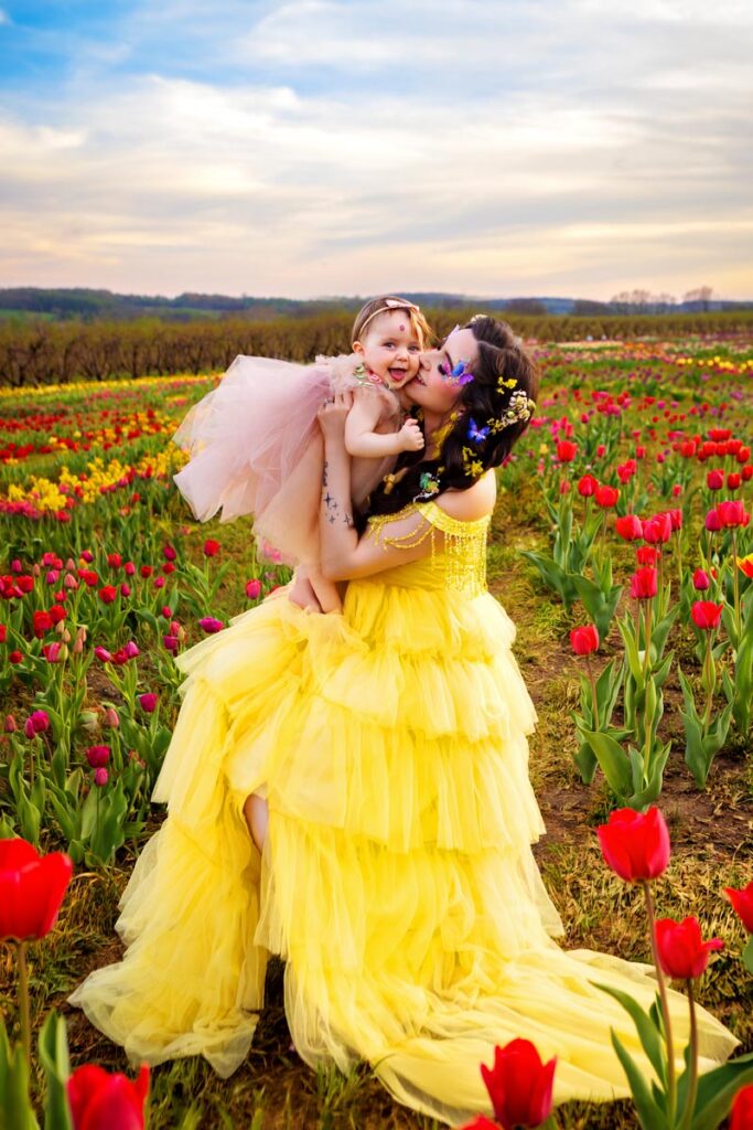 A woman in a flowing yellow dress lifts and kisses a smiling young girl in a pink dress, surrounded by blooming red tulips in a vibrant field under a partly cloudy sky at sunset.