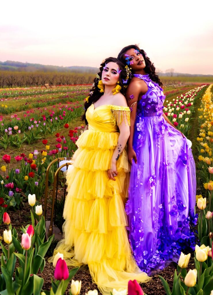 Two women in bright, flowing dresses (purple, blue, yellow, green) pose on a tulip field at dusk, with dramatic lighting and a vibrant sunset sky in the background.