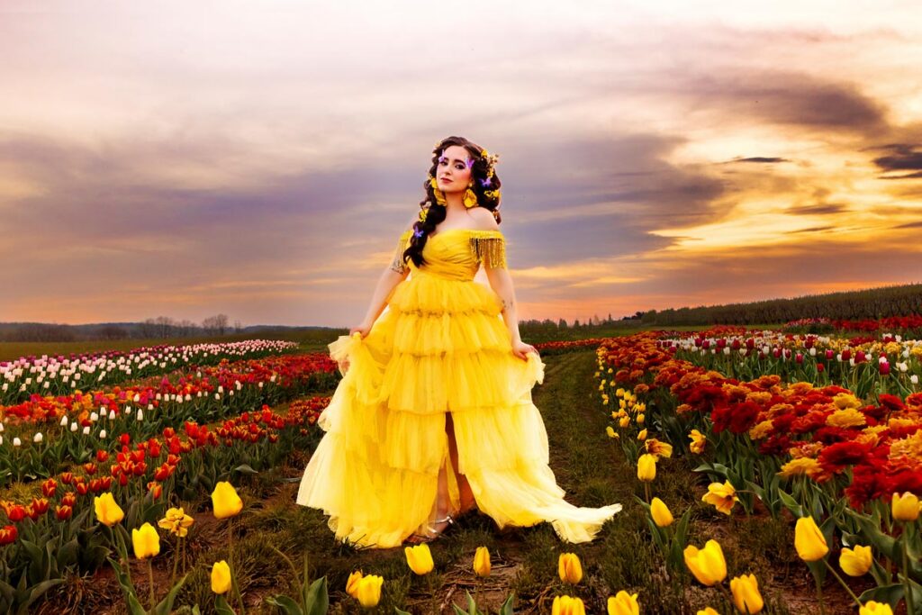 A woman in a flowing yellow dress stands in a vibrant field of red, orange, and yellow tulips under a cloudy sky, with green hills in the distance.