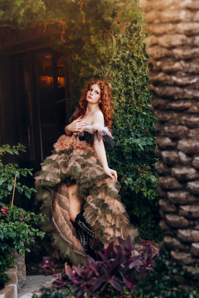 A woman with long curly red hair poses outdoors in a ruffled, off-shoulder gown surrounded by lush greenery and plants, with sunlight filtering through the background.