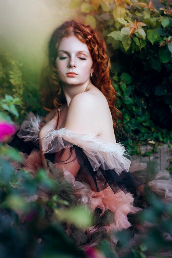 A woman with long red hair and fair skin poses outdoors among green foliage, wearing a ruffled, off-the-shoulder gown. She looks downward with a serene expression, surrounded by blurred flowers and leaves.
