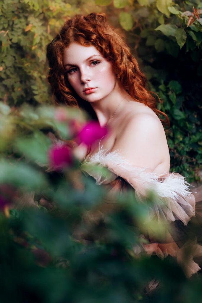 A young woman with long, curly red hair sits outdoors surrounded by lush greenery and blurred pink flowers, gazing softly at the camera with a serene expression.