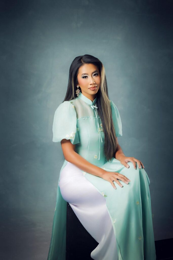A woman with long hair wears a light green traditional dress from her heritage with sheer sleeves and white pants, sitting on a stool against a gray studio backdrop.
