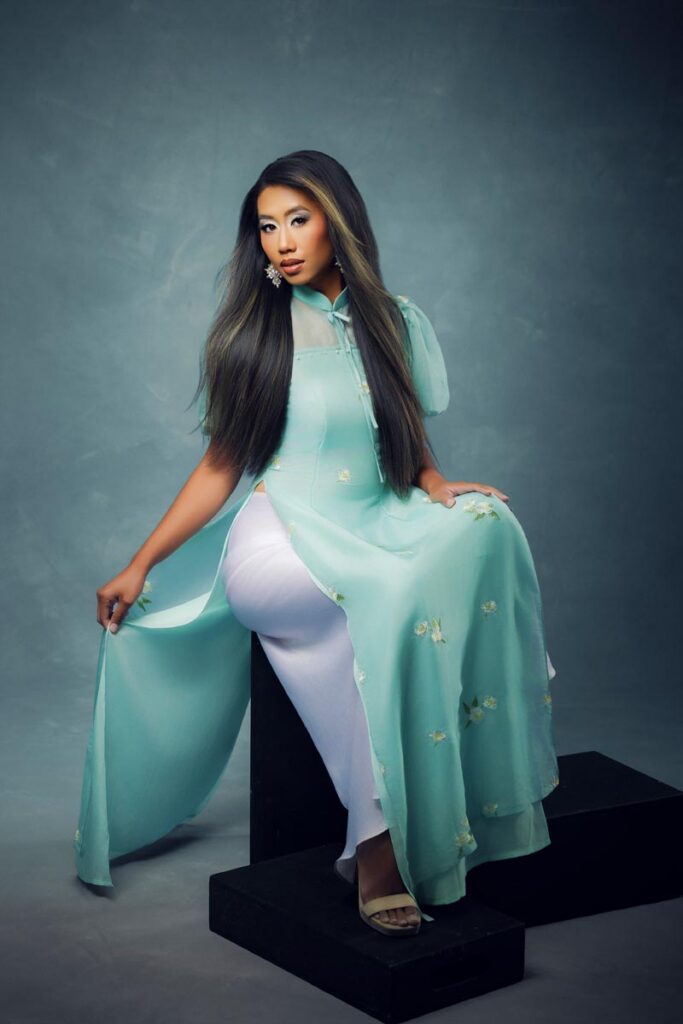 A woman with long dark hair sits on a black block, wearing a light blue, embroidered áo dài dress over white pants, posing against a gray studio backdrop.