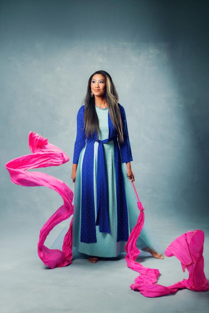 A woman in a long blue and teal dress stands barefoot on a gray floor, holding flowing pink fabric that swirls around her as part of her heritage, against a gray background.