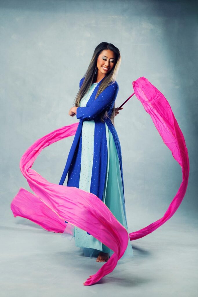 A woman in a blue and light green dress dances gracefully, holding bright pink silk ribbons that swirl around her against a plain gray background.