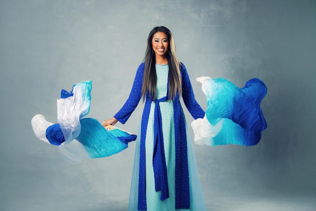 A woman in a long blue dress smiles while waving two blue and white flowing scarves against a light gray background.
