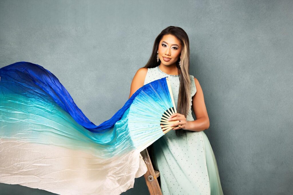 A woman in a light blue dress holds a large fan with blue, teal, and white flowing fabric against a gray background.
