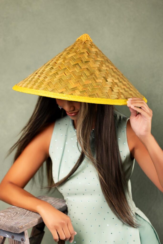 A woman in a pale green dress sits on a wooden stool, tilting a woven conical hat over her face as part of her heritage, with long dark hair falling forward against a green background.