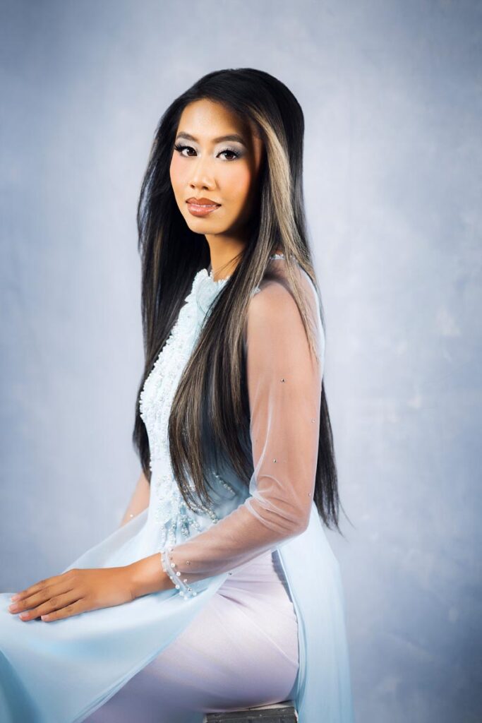 Woman with long, straight dark hair in a sheer, light blue traditional dress poses seated against a soft, light blue background, looking confidently at the camera.