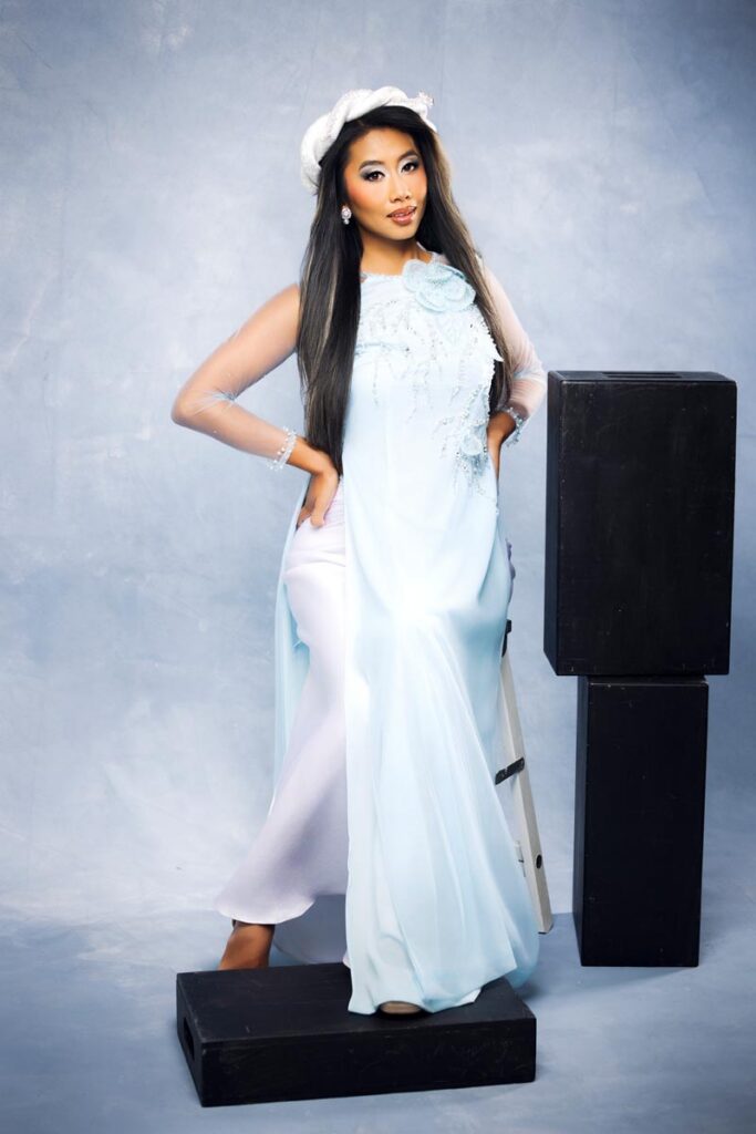 A woman in a light blue dress and white beret poses confidently beside black blocks, with one hand on her hip and the other resting on the blocks, against a soft blue background.