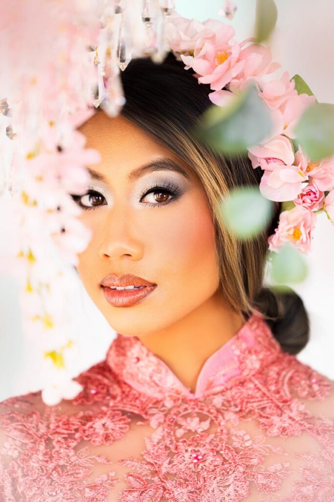 A woman in a pink lace dress and floral headpiece gazes at the camera, surrounded by soft, pink flowers and greenery, with bright, natural light illuminating her face and heritage.