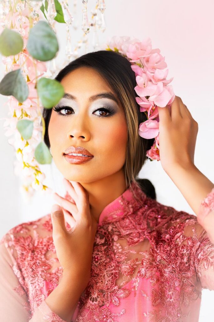 A woman in a pink lace dress and pink flower crown poses gracefully, touching her face and head. She has long dark hair, glossy lips, and soft makeup, with greenery and soft lighting in the background.