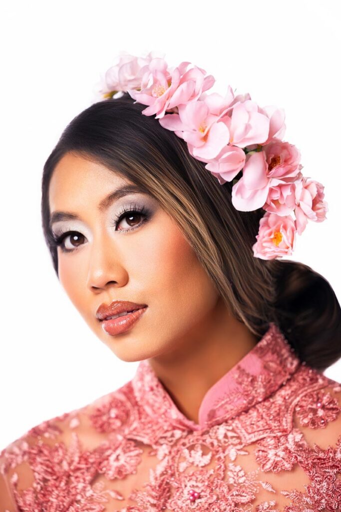 Woman wearing a pink floral headpiece and embroidered pink dress, with smooth makeup and long hair styled back, looking slightly to the side against a white background.