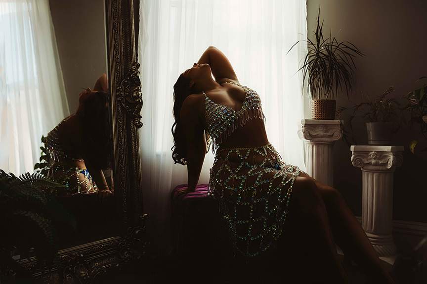 A person wearing jeweled lingerie with gold chains and crystal accents poses near a mirror, showcasing the intricate details and reflections of the sparkling costume.
