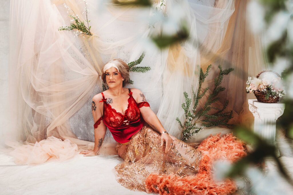 A person with short light hair wears a red top and a flowing gold and orange skirt. They pose gracefully in front of soft, sheer drapes with green foliage, creating a dreamy, ethereal atmosphere.